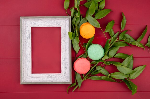 Bovenaanzicht van wit frame met kleurrijke macarons en bladtakken op een rood oppervlak