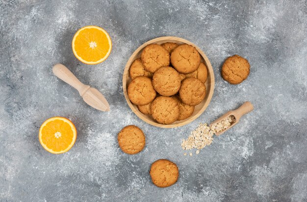 Bovenaanzicht van zelfgemaakte koekjes op een houten bord en havermout met sinaasappelen over grijze ondergrond.