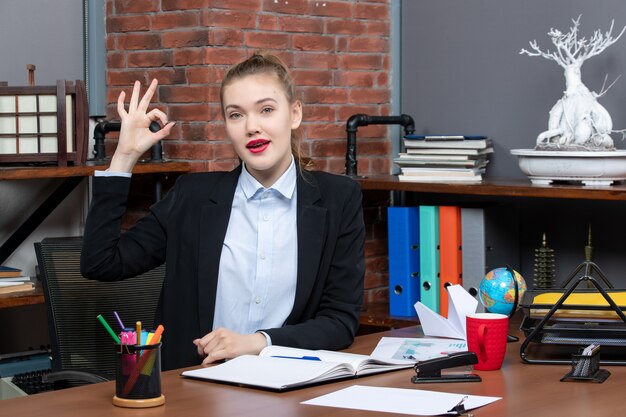 Bovenaanzicht van zelfverzekerde jonge vrouw die aan een tafel zit en het document vasthoudt en een brilgebaar maakt op kantoor