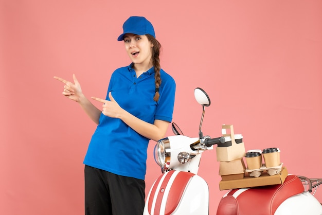 Bovenaanzicht van zelfverzekerde koeriersdame die naast de motorfiets staat met koffie en kleine taarten erop op een pastelkleurige perzikkleurige achtergrond