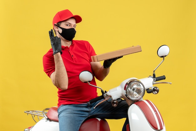 Bovenaanzicht van zelfverzekerde tevreden koeriersmens die rode blouse en mutshandschoenen draagt in medisch masker zittend op een scooter met twee wachtrij