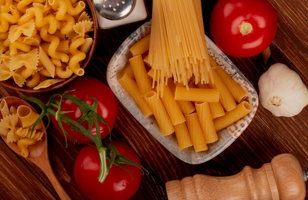 Bovenaanzicht van ziti pasta in kom met spaghetti en andere soorten in kom en lepel zout tomaten knoflook op houten oppervlak