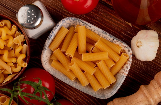 Bovenaanzicht van ziti pasta met verschillende soorten in kom en zout tomaten knoflook op houten oppervlak