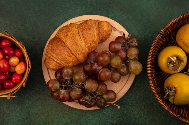 Bovenaanzicht van zoete druiven op een houten keukenbord met croissant met kaki fruit op een emmer op een groen oppervlak