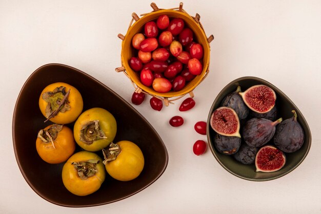 Bovenaanzicht van zoete zwarte vijgen op een kom met cornelian kersen op een emmer met kaki fruit op een kom op een witte muur