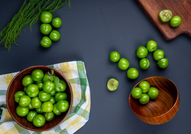 Gratis foto bovenaanzicht van zure groene pruimen in houten kommen en houten snijplank met gesneden pruimen op zwarte tafel