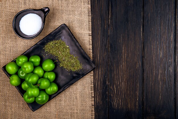 Bovenaanzicht van zure groene pruimen met gedroogde pepermunt op een zwart dienblad en zout op zak op donkere houten tafel met kopie ruimte