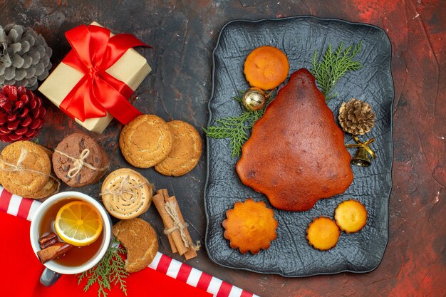 Bovenaanzicht verschillende koekjes vastgebonden met touw kopje thee kaneelstokjes cadeau taarten op zwarte schotel op donkerrode tafel