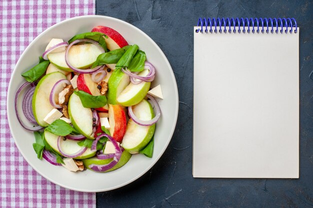 Bovenaanzicht verse appelsalade in kom paars wit geruit tafelkleed notitieblok op donkere tafel