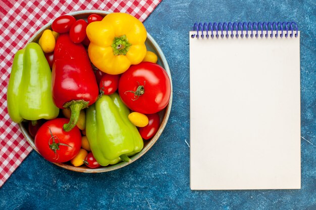 Bovenaanzicht verse groenten kerstomaatjes verschillende kleuren paprika tomaten cumcuat op plaat op rood en wit geruit tafelkleed Kladblok op blauw oppervlak