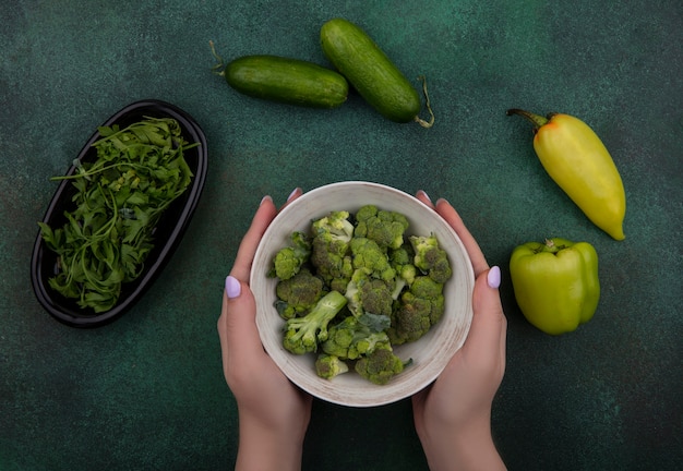 Bovenaanzicht vrouw met broccoli in een kom met komkommers kruiden en paprika's op een groene achtergrond