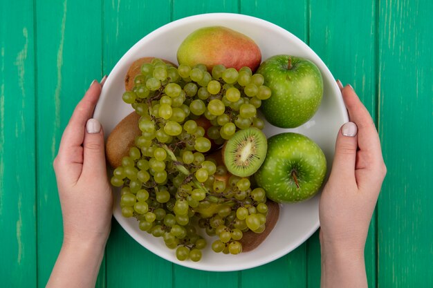 Bovenaanzicht vrouw met groene appels met kiwi groene druiven en peer op een plaat op een groene achtergrond