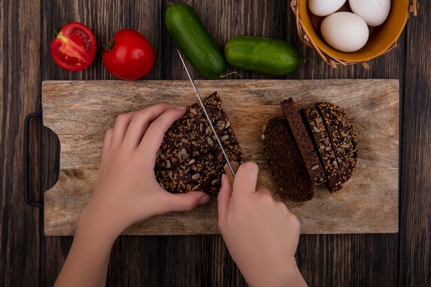 Bovenaanzicht vrouw snijdt zwart brood op een stand met tomaten, komkommers en kippeneieren op een houten achtergrond