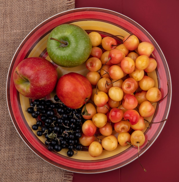 Bovenaanzicht witte kers met zwarte bessen perzik en gekleurde appels op een bord op een rode tafel