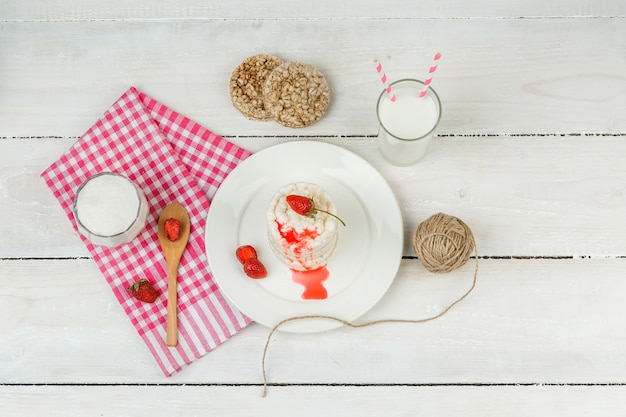 Bovenaanzicht witte rijstwafels en aardbeien op plaat met rood geruit tafelkleed, houten lepel en zuivelproducten op witte houten plank oppervlak. horizontaal