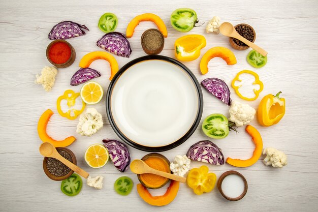 Bovenaanzicht witte schotel gesneden groenten rode kool pompoen bloemkool gele paprika kruiden in kleine kommen op witte houten tafel