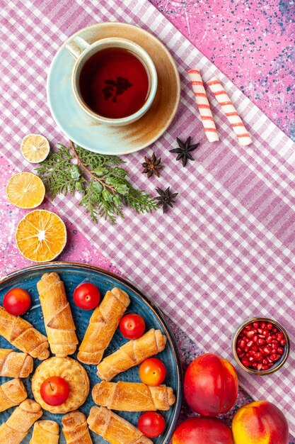 Bovenaanzicht zoete heerlijke bagels in dienblad met pruimen, verse perziken en kopje thee op lichtroze bureau