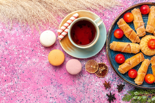 Bovenaanzicht zoete heerlijke bagels in dienblad met verse zure pruimen franse macarons en kopje thee op lichtroze bureau