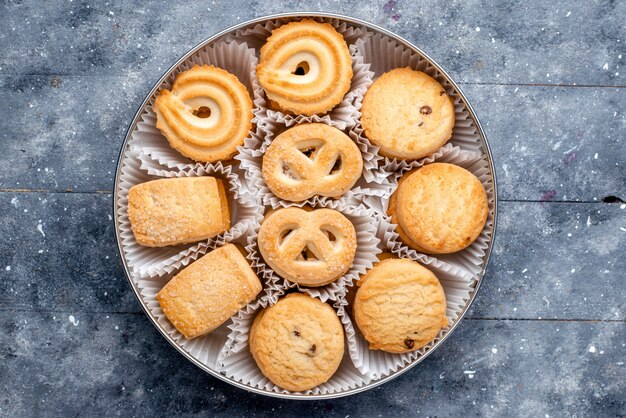 Bovenaanzicht zoete heerlijke koekjes verschillend gevormd binnen rond pakket op het grijze koekjeskoekje van de bureausuiker zoete cake