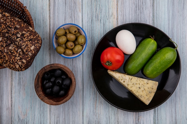 Bovenaanzicht zwart brood met kaas komkommers tomaat en ei op een bord met zwarte en groene olijven op grijze achtergrond