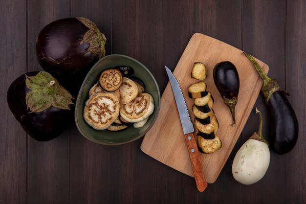 Bovenaanzicht zwarte aubergine met wiggen in een kom en op een snijplank met een mes op een houten achtergrond