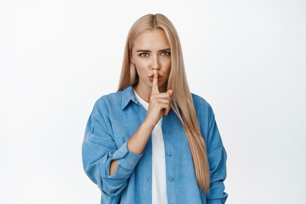 Boze blonde vrouw zwijgt, maakt shhh-geluid en kijkt teleurgesteld, zegt dat ze stil moet zijn, zwijg alsjeblieft, zwijg gebaar, staande op wit.