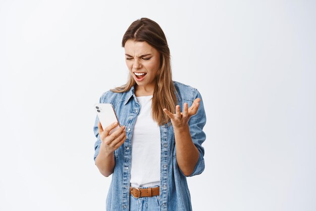 Boze en geërgerde jonge vrouw die pissig naar de mobiele telefoon staart en klaagt over het zien van slecht nieuws op het smartphonescherm dat tegen een witte achtergrond staat