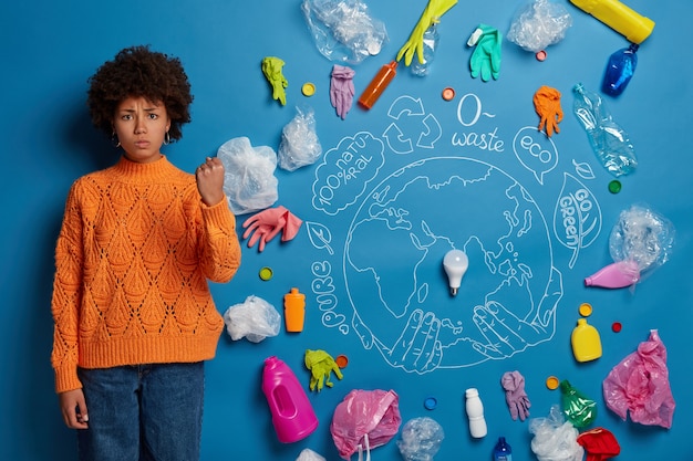 Gratis foto boze geïrriteerde vrouw vraagt om vervuiling te vermijden, toont vuist, eist van mensen die ecologisch vriendelijk zijn, heeft een strikte blik, getekende planeet en veel plastic afval dat een ernstig probleem aantoont