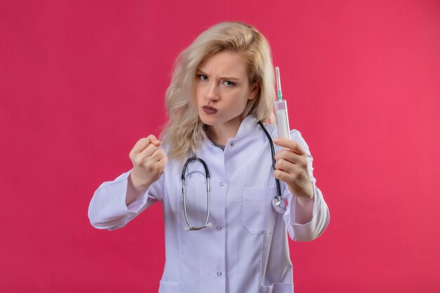 Boze jonge arts die stethoscoop in de spuit van de medische togaholding op isolatie rode backgroung draagt