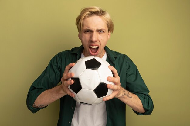 Boze jonge blonde kerel die de groene bal van de t-shirtholding draagt