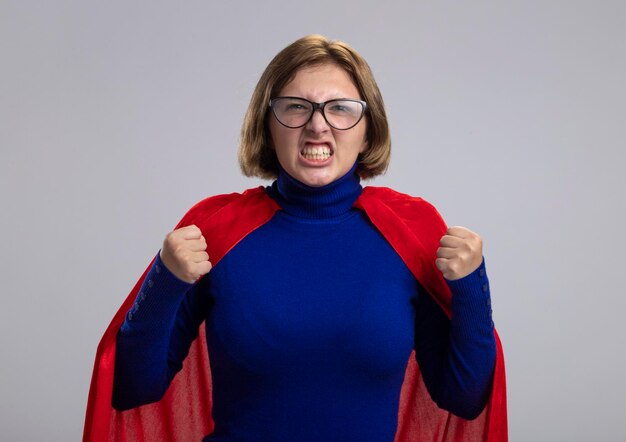 Boze jonge blonde superherovrouw in rode cape die glazen draagt die voorste gebalde vuisten bekijken die op witte muur worden geïsoleerd