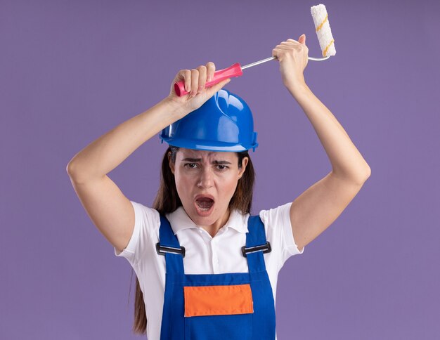 Boze jonge bouwersvrouwen in uniform die miniverfroller opheffen die op purpere muur wordt geïsoleerd