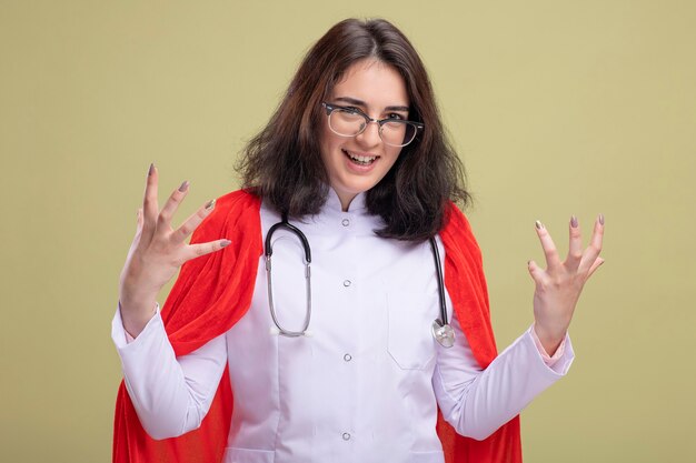 Boze jonge Kaukasische superheld meisje in rode cape dragen dokter uniform en stethoscoop met bril houden handen in de lucht geïsoleerd op olijf groene muur