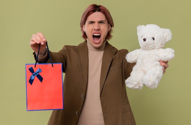 Boze jonge knappe man met witte teddybeer en cadeautas die tegen iemand schreeuwt