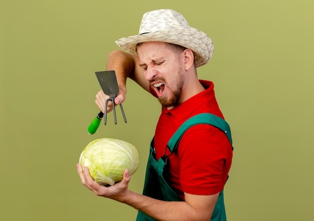 Boze jonge knappe Slavische tuinman in uniform en hoed staande in profielweergave houden kijken naar kool houden schoffel-hark erboven in een andere hand geïsoleerd