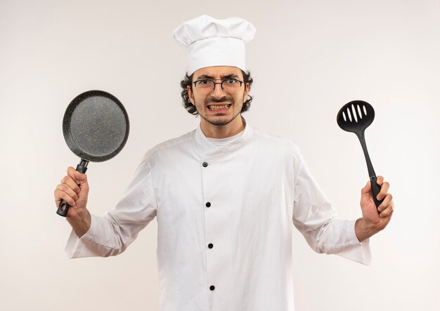 Boze jonge mannelijke kok die eenvormige chef-kok en glazen draagt die pan en spatel houden