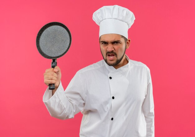 Boze jonge mannelijke kok in uniform van de chef-kok met koekenpan geïsoleerd op roze muur