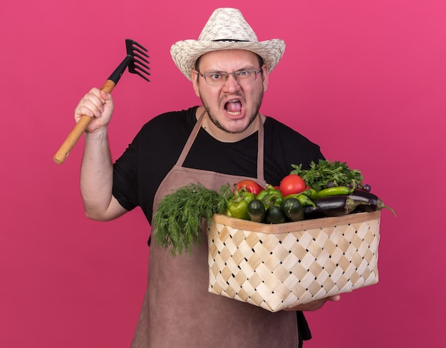 Gratis foto boze jonge mannelijke tuinman die het tuinieren hoed draagt die plantaardige mand houdt die hark opheft die op roze muur wordt geïsoleerd