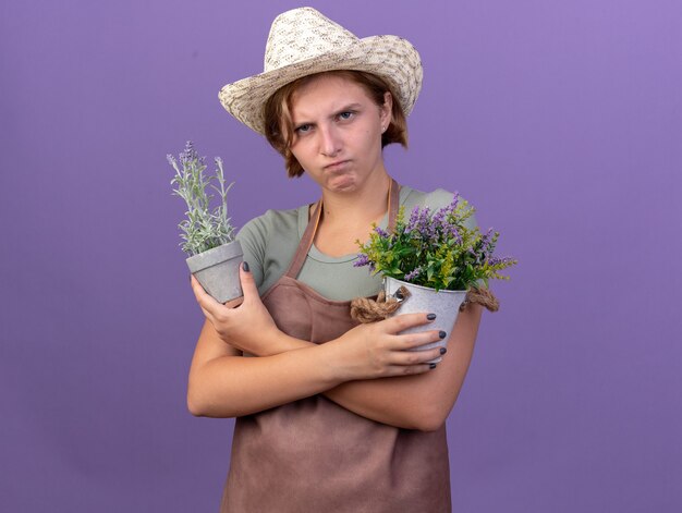 Boze jonge Slavische vrouwelijke tuinman die tuinieren hoed draagt die bloemen in bloempotten op paars houdt