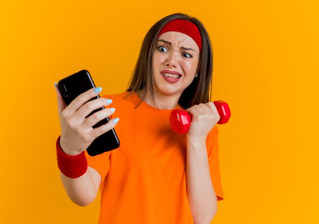 Boze jonge sportieve vrouw die hoofdband en polsbandjes draagt die domoor en mobiele telefoon houden en mobiele telefoon bekijken