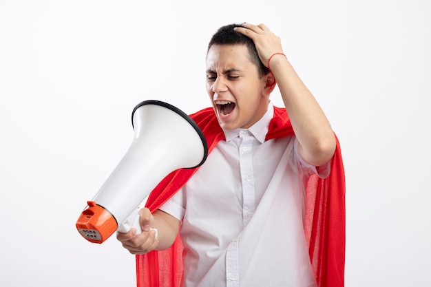 Boze jonge superheld jongen in rode cape houden en kijken naar spreker hand zetten hoofd schreeuwen geïsoleerd op een witte achtergrond