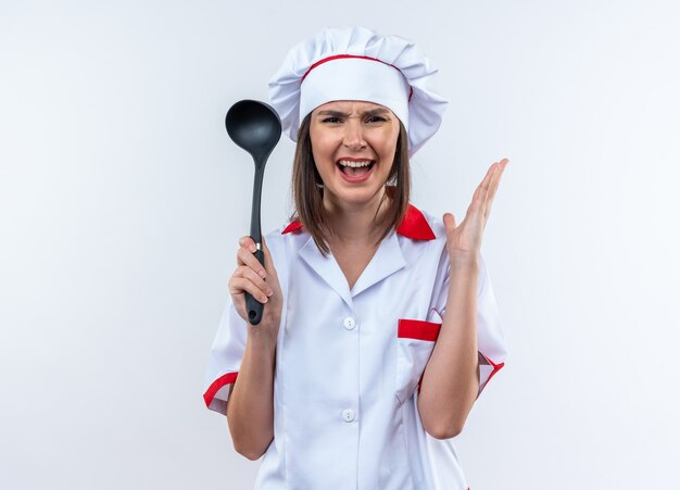 Boze jonge vrouwelijke kok die chef-kok uniform draagt die pollepel houdt die op witte achtergrond wordt geïsoleerd