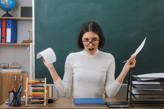 boze jonge vrouwelijke leraar die een bril draagt, scheurt papier aan het bureau met schoolhulpmiddelen in de klas