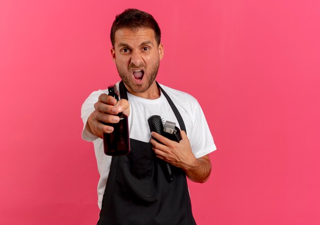 Boze kapper man in schort met haarborstel en trimmer gericht naar voren met spray staande over roze muur