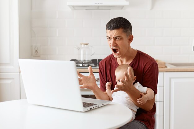 Boze knappe man met donker haar met een bordeauxrood casual t-shirt, aan het werk op een laptop tijdens het babysitten, kijkend naar het display met agressieve blik, schreeuwend, poserend in de witte keuken.