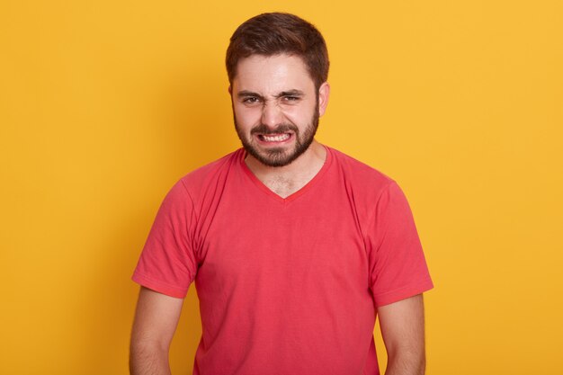 boze man met rode casual t-shirt, ontevreden, houdt handen gespannen, ongeschoren man met stijlvol kapsel verdraaide zijn gezicht met woede, poseerde geïsoleerd op geel.