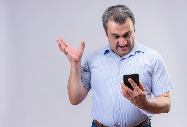 Boze man van middelbare leeftijd in blauw verticaal gestreept overhemd die zijn mobiele telefoon bekijkt terwijl hij staat