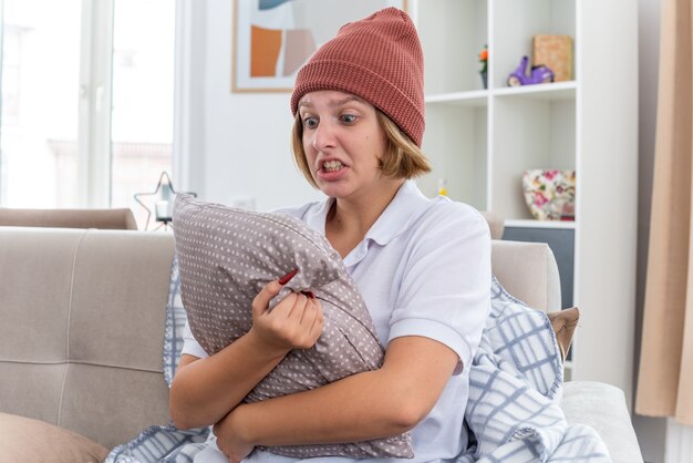 Boze ongezonde jonge vrouw in warme muts met deken die er onwel en ziek uitziet en lijdt aan verkoudheid en griep met kussen met agressieve uitdrukking zittend op de bank in lichte woonkamer