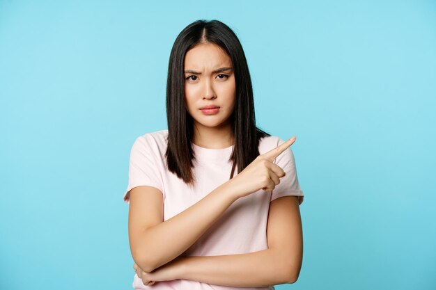 Boze ontevreden aziatische vrouw wijzend naar rechts gefronste wenkbrauwen in afkeuring staande in t-shirt over b...