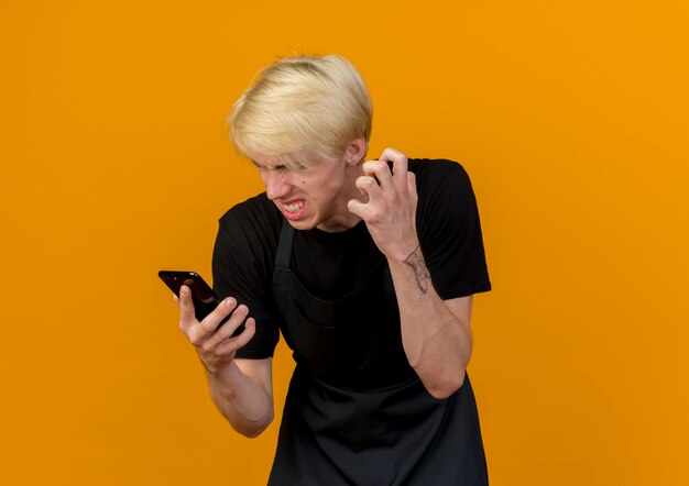 Boze professionele kapper man in schort kijken naar zijn smartphonescherm gaan wild staande over oranje muur